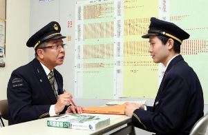 改札業務についての問題集をもとに、吉川直輝さん（右）を指導する沢正和さん。後ろには進み具合を示す一覧表が貼られている＝ＪＲ名古屋駅で